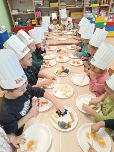 Przedszkolaki kroją owoce przy stolikach.