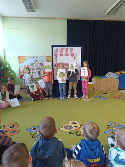 Uczniowie stoją i trzymają w rękach ilustracje z warzywami.