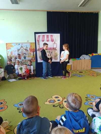 Uczniowie stoją i trzymają w rękach ilustracje z warzywami.