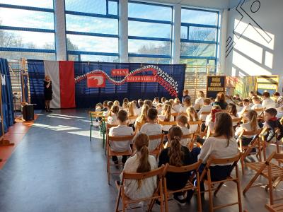 Dyrekcja przemawia do uczniów, którzy siedzą na krzesłach.