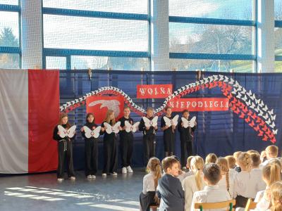 Uczniowie ubrani na czarno stoją na scenie i trzymają w dłoniach białe motyle z literami. Litery tworzą napis: 