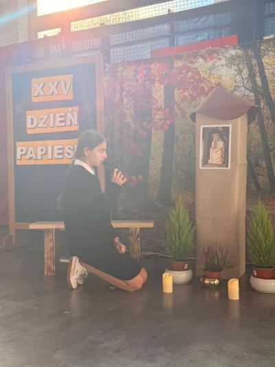 Uczennica klęczy i mówi wiersz w kierunku kapliczki z Panem Jezusem Frasobliwym.