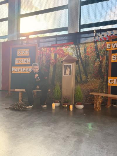 Uczeń siedzi na drewnianej ławeczce i mówi wiersz w kierunku kapliczki z Panem Jezusem Frasobliwym.