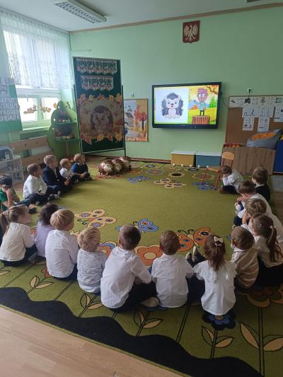 Dzieci siedzą w kole na dywanie i oglądają film edukacyjny o jeżu.