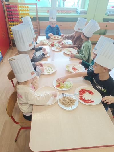 Dzieci w czapkach kucharskich nakładają warzywa i kurczaka na tortille.