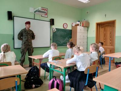 Żołnierz stoi pod tablicą w klasie i opowiada, a dzieci siedzą w ławkach i aktywnie go słuchają.