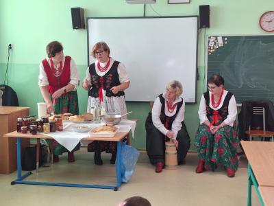 Jedna pani z Koła Gospodyń Wiejskich opowiada, a druga robi masło w maślniczce. Dwie pozostałe im towarzyszą