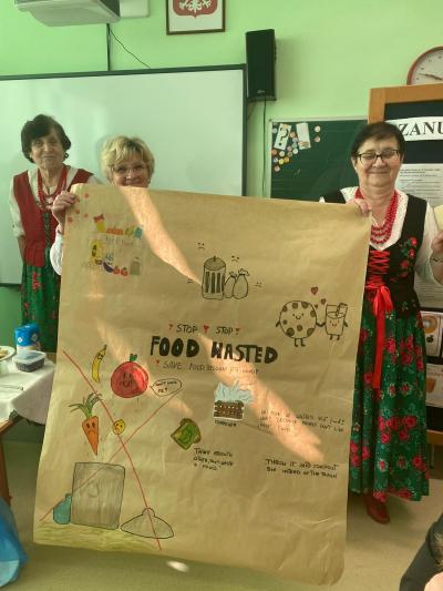 Panie z Koła Gospodyń Wiejskich trzymają plakat wykonany przez uczniów na temat marnowania żywności.