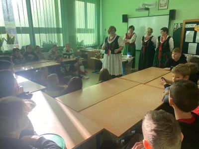 Dzieci siedzą w ławkach ułożonych w podkowę i słuchają opowiadania Pań z Koła Gospodyń Wiejskich.