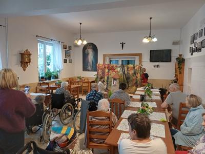 Uczennica klęczy obok kapliczki i mówi swoją kwestię do mikrofonu, a osoby chore i starsze słuchają.