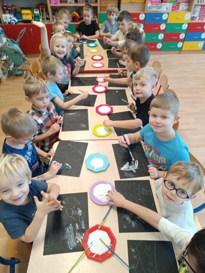 Przedszkolaki malują solą na czarnych kartkach.
