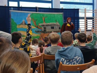 Uczniowie siedzą na krzesłach i oglądają spektakl. Na scenie stoi aktor przebrany za Misia.