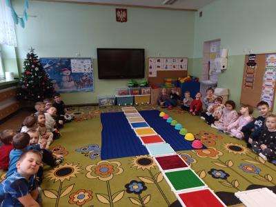 Dzieci siedzą na dywanie. Na środku dywanu rozłożone są dwie ścieżki sensoryczne.