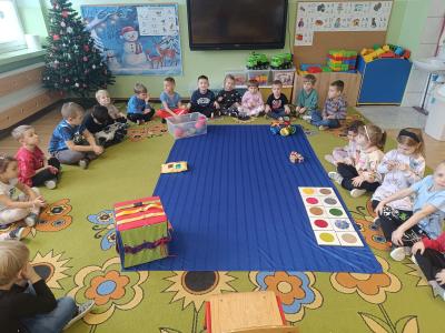 Dzieci siedzą w kole na dywanie. W środku koła znajdują się pomoce sensoryczne.