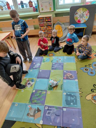 Dzieci siedzą w kole na dywanie, a jedno dziecko programuje trasę robota Photon po macie edukacyjnej.