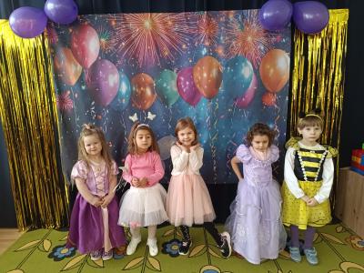 Dzieci przebrane za księżniczki i pszczółkę stoją na karnawałowym tle z balonami.