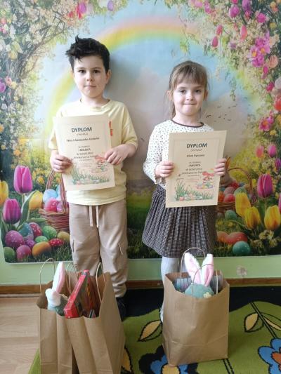 Dzieci trzymają dyplomy, a przed nimi stoją torebki z prezentami.