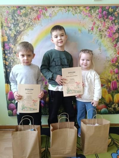 Dzieci trzymają dyplomy, a przed nimi stoją torebki z prezentami.