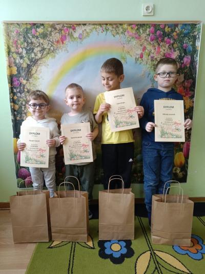 Dzieci trzymają dyplomy, a przed nimi stoją torebki z prezentami.