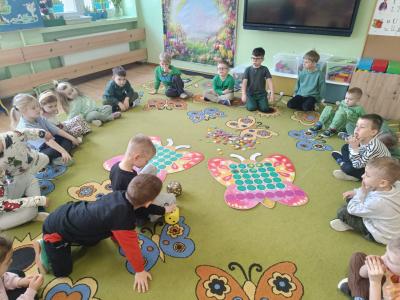 Dzieci siedzą w kole na dywanie, a po środku nich znajdują się dwa duże motyle i kostki do gry.