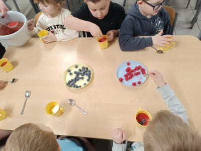 Dzieci się przy stołach, na których są kubki na jogurt, owoce i mus.