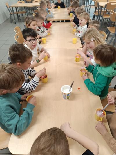 Dzieci się przy stołach, na których są kubki na jogurt, owoce i mus.