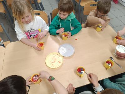 Dzieci się przy stołach, na których są kubki na jogurt, owoce i mus.