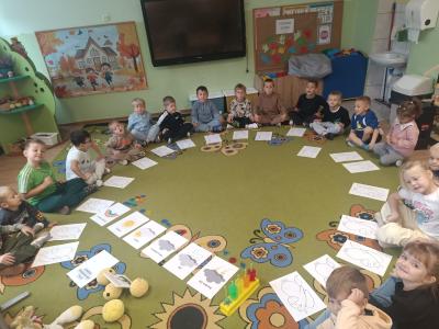 Dzieci siedzą w kole na dywanie, a przed nimi znajdują się karty pracy.
