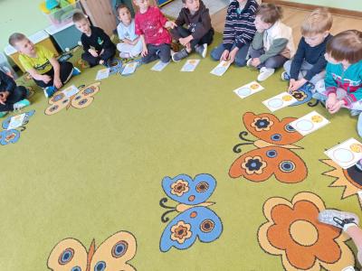 Dzieci siedzą w kole na dywanie, a przed nimi znajdują się karty pracy-radość.